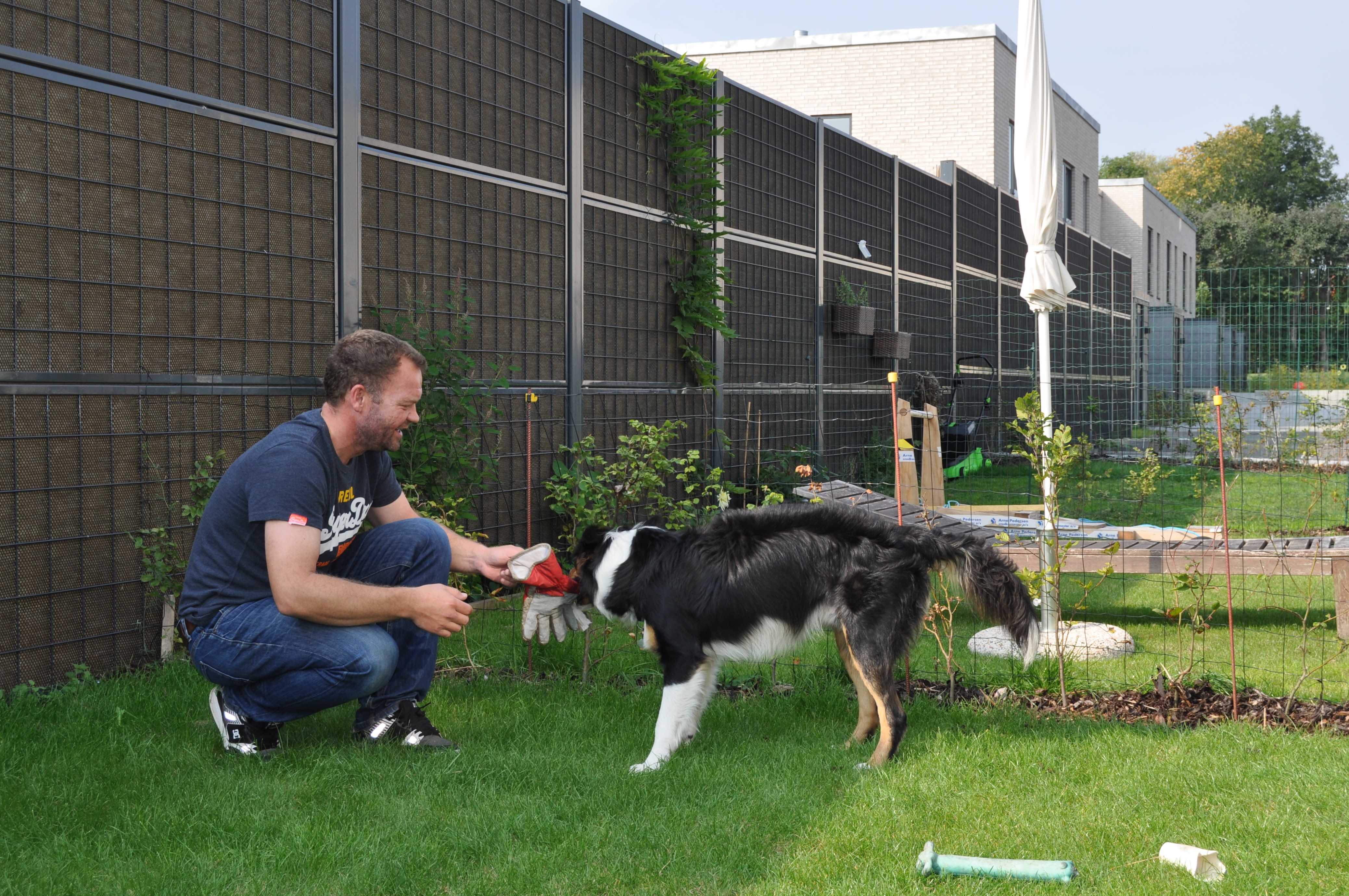 En mand og hans hund sidder foran et Noistop® Steel støjhegn fra Poda Hegn der er opsat i en have til afskærmning af støj fra naboerne